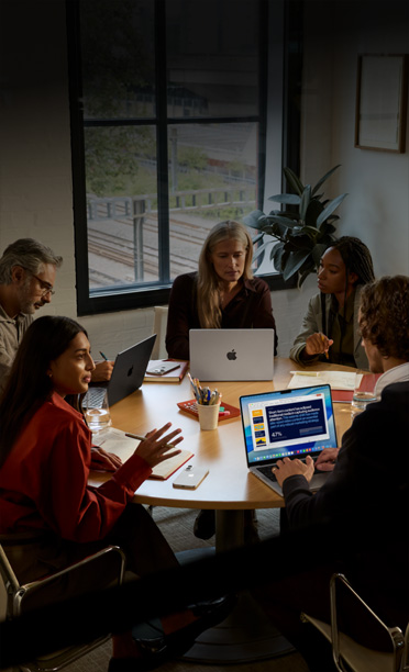 Cinco pessoas sentadas ao redor de uma mesa de reunião, colaborando em notebooks MacBook, com aparelhos iPhone virados para baixo sobre a mesa