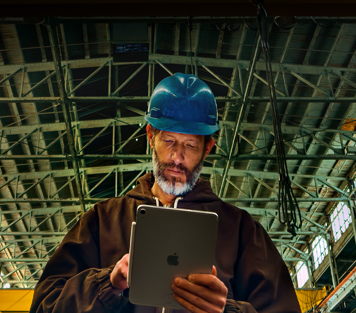 iPad prateado com um Apple Pencil fixado, segurado por uma pessoa com uma jaqueta marrom e capacete azul trabalhando em um ambiente industrial com vigas metálicas no teto