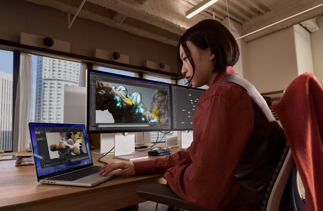 Uma pessoa trabalha em uma mesa com um MacBook Pro conectado a um monitor externo. As duas telas mostram uma renderização 3D, com edifícios visíveis pela janela ao fundo