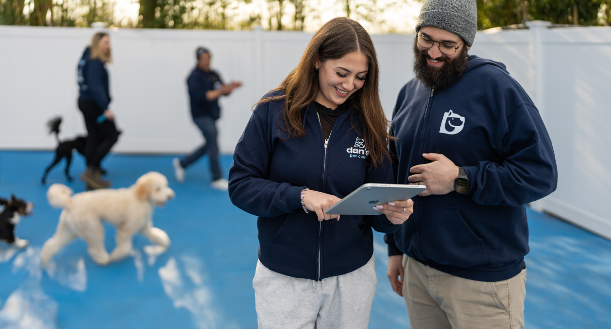 Dois funcionários usando moletons com a marca da empresa olham juntos para um iPad em uma área de recreação para cães, com cães e outros funcionários ao fundo