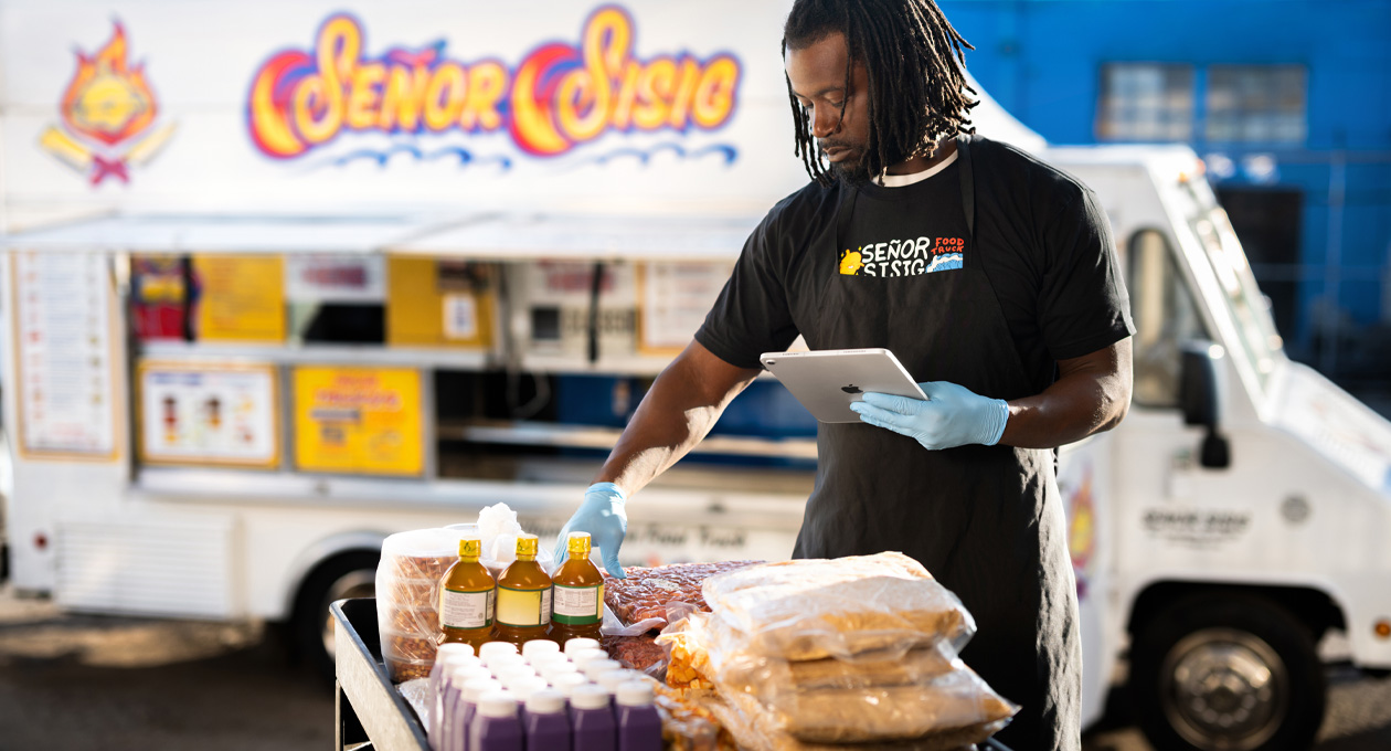 Uma pessoa usando camiseta da marca e luvas consulta um iPad enquanto prepara alimentos sobre uma mesa, com um food truck ao fundo