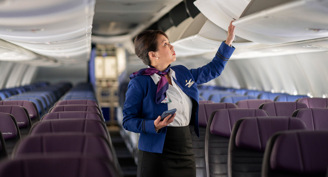 Uma comissária de bordo segura um iPhone enquanto abre um compartimento superior de bagagem em uma cabine de avião vazia