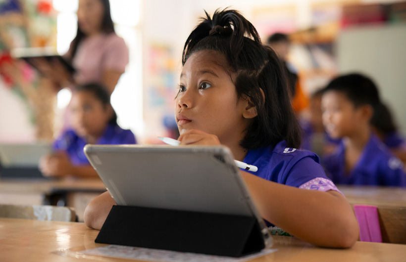 A young girl sits at her desk, looking towards the front of the classroom and is fully engaged. She is using an iPad and holding an Apple Pencil.