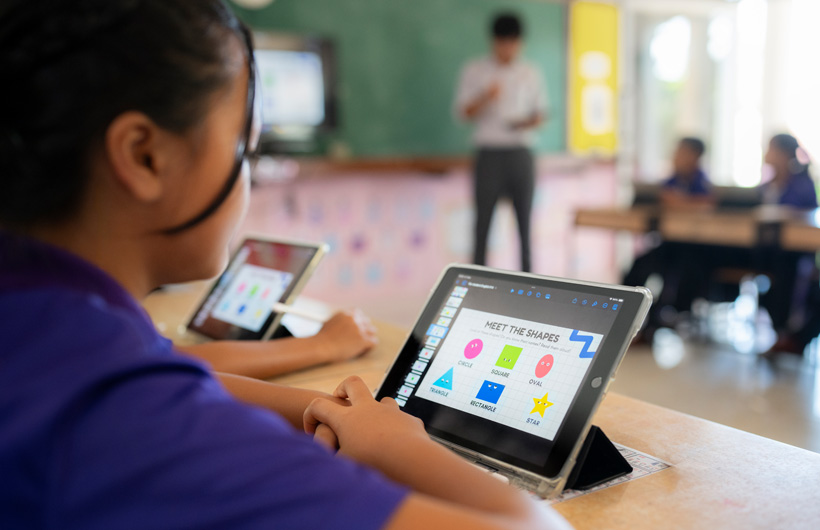 Close-up of a student at their desk, using an iPad. On screen, there are colourful shapes displayed with the title, ‘Meet the shapes’.