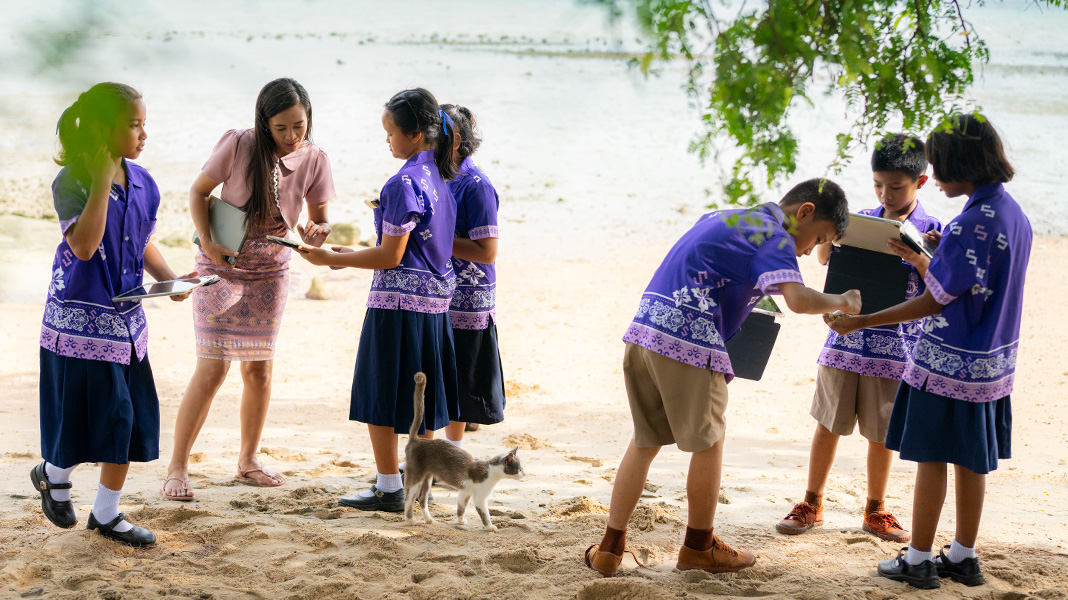 Six elementary students in colourful purple uniforms stand on a sandy beach holding iPads. A teacher is assisting one student on an iPad, and there is a cat walking among them.
