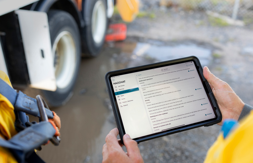 Bàn tay cầm iPad đang mở ứng dụng HazChat, hiển thị các mục Chi Tiết Công Việc, Lập Kế Hoạch, Rủi Ro và Biện Pháp Kiểm Soát, trong khi một người đang đứng tại công trường bên vũng bùn