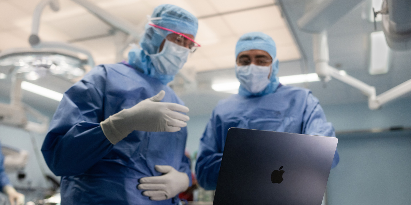 수술실에 있는 두 명의 외과의가 MacBook을 보며, 한 명은 손으로 손짓을 하고 다른 한 명은 지켜보고 있는 모습