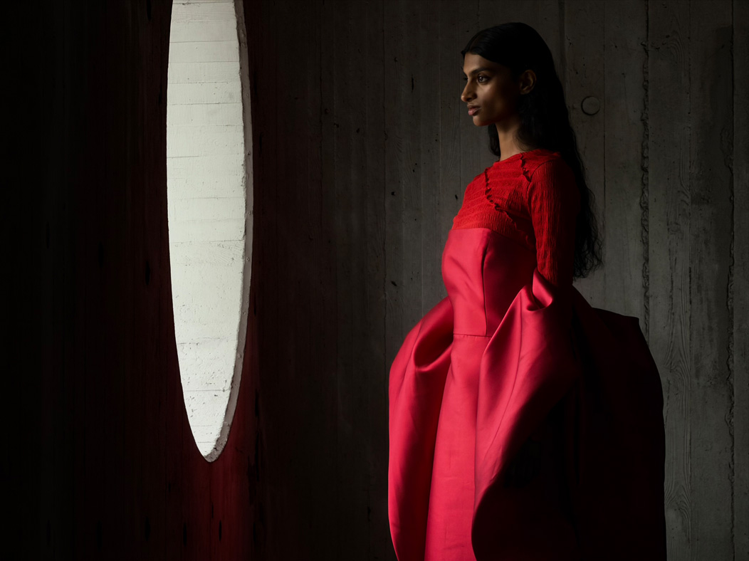 Una mujer con vestido largo rojo está siendo fotografiada de pie en una sala vacía. La distancia focal de la imagen avanza en la app Fotos para demostrar las capacidades del sistema de cámaras Pro, se muestra una distancia focal de 200 mm